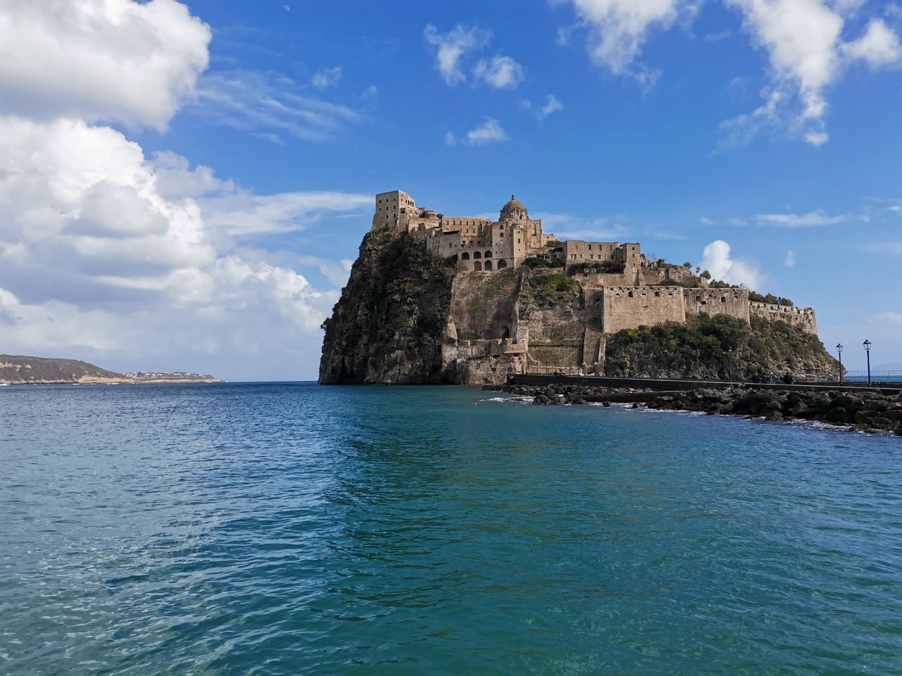 The Aragonese Castle of Ischia, a fortress on a tall rocky islet above the blue sea, seen from the shore under a cloudy sky.