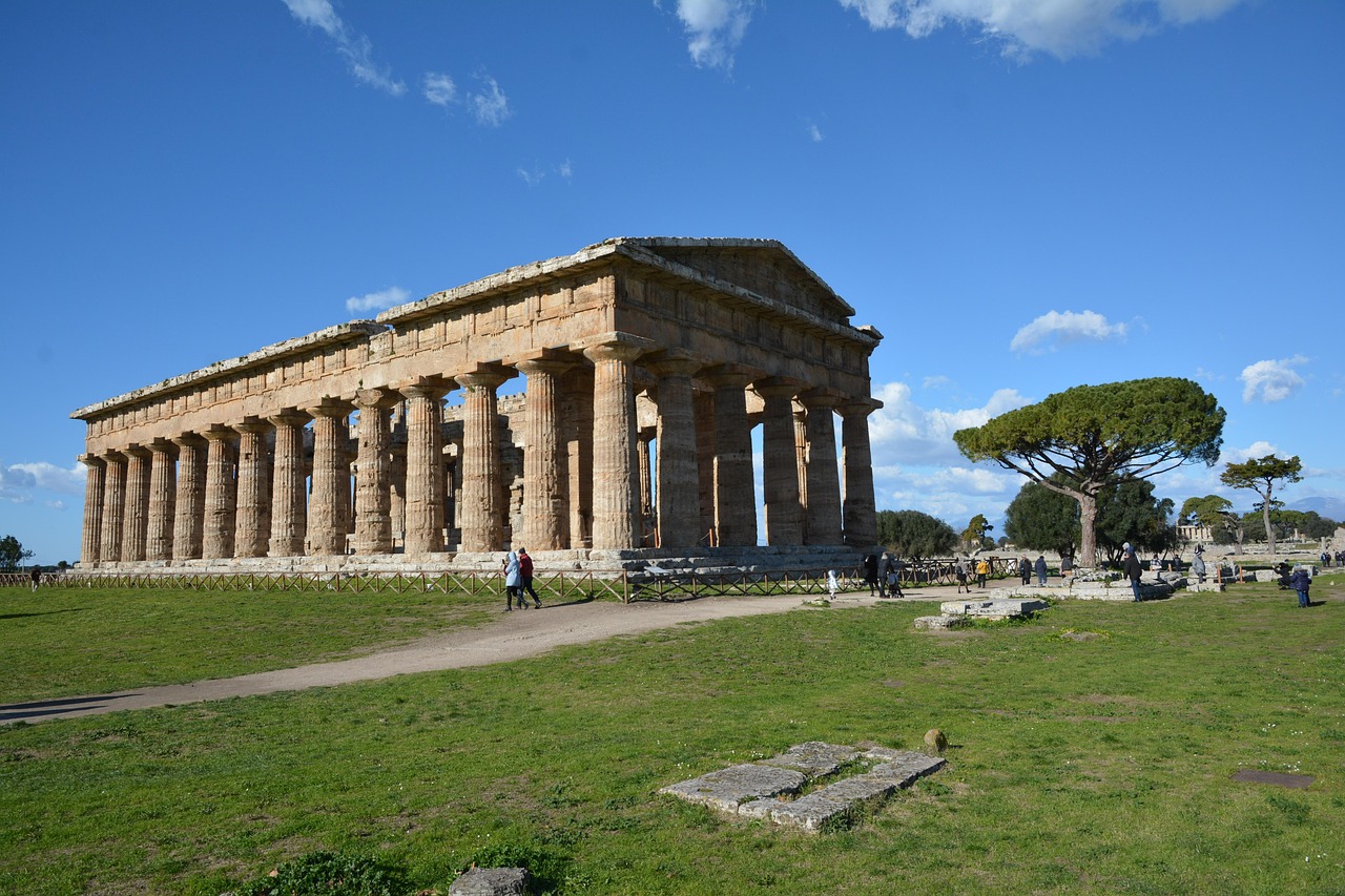 The Temple of Neptune at Paestum, a large Doric temple with a row of stone columns, seen across a grassy lawn under a blue sky.
